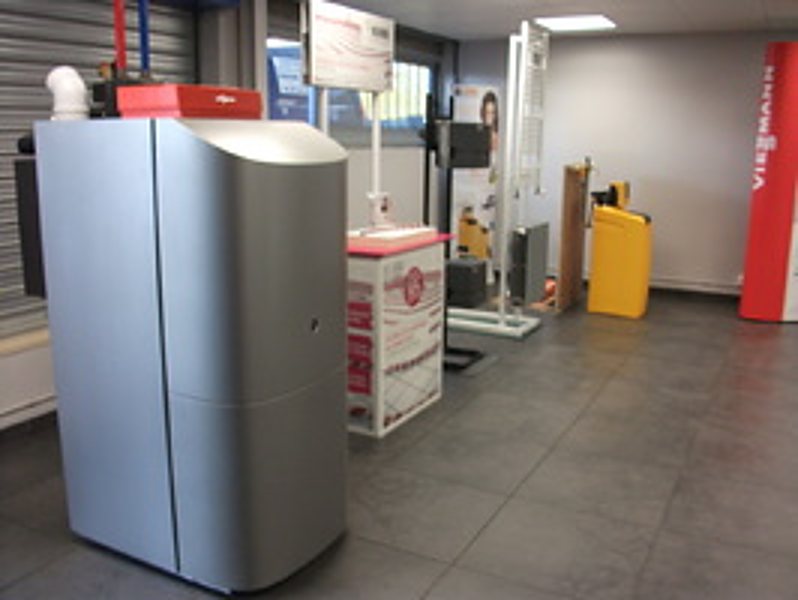 Une grande chaudière grise à côté de présentoirs et d'appareils de chauffage dans un showroom au sol carrelé gris.