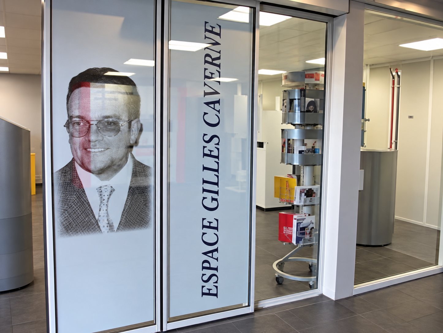 Vitrine d'un local orné d'un grand portrait d'homme en costume et du texte vertical « Espace Gilles Caverne », avec des présentoirs visibles à l'intérieur.
